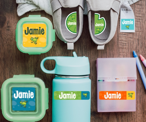 Assorted items for children, including two plastic storage containers, a water bottle, a crayon box, a pair of slip-on shoes, and a towel, all labeled with stick-on labels that have frogs and the name Jamie on them.