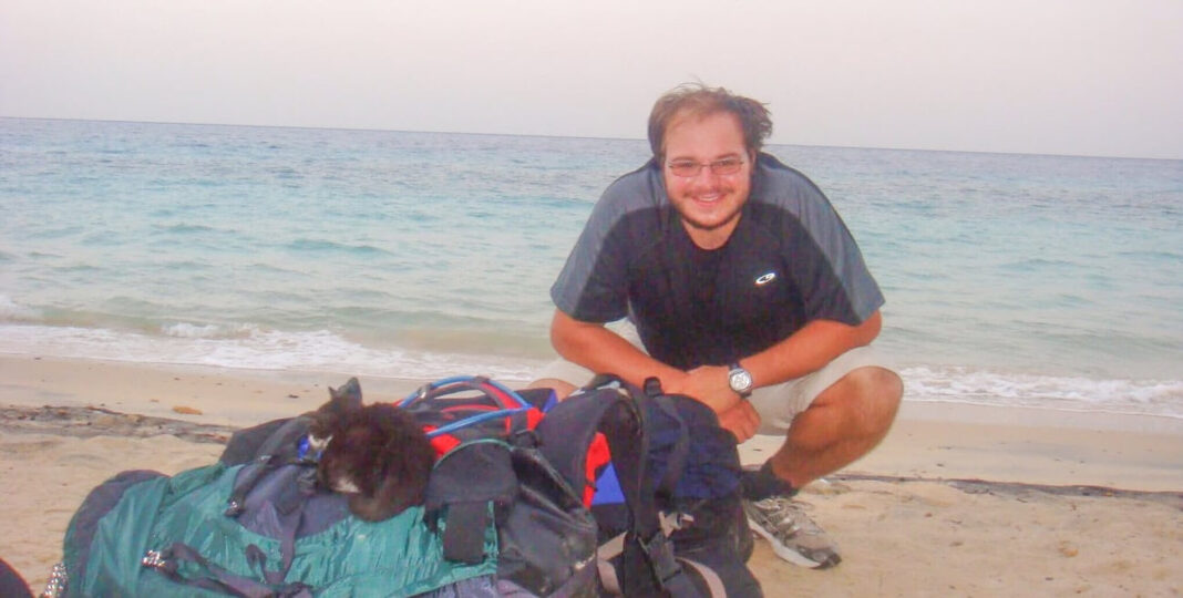 a man on a beach crouched behind two backpacks with a black kitten curled up on one of them