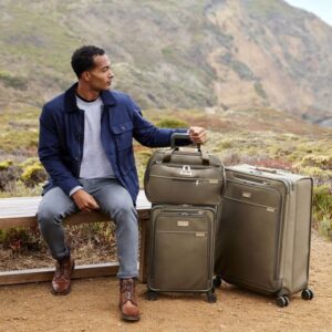 A man sits on a bench in front of a mountain in front of a field with three olive green suitcases next to him from sustainable luggage brand Briggs & Riley.