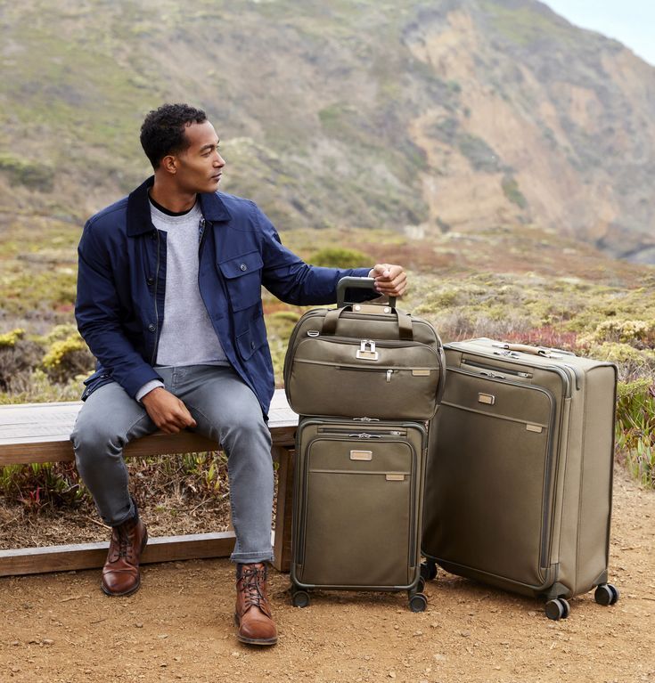 A man sits on a bench in front of a mountain in front of a field with three olive green suitcases next to him from sustainable luggage brand Briggs & Riley.