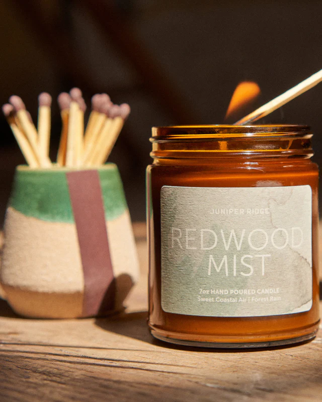 A candle being lit. The candle is in a brown jar and has the words Redwood Mist and Juniper Ridge on it.