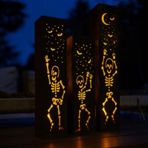 Three solar-powered metal luminary pillars with dancing skeletons carved on them are lit from within, made in America by Northern Forge, LLC.