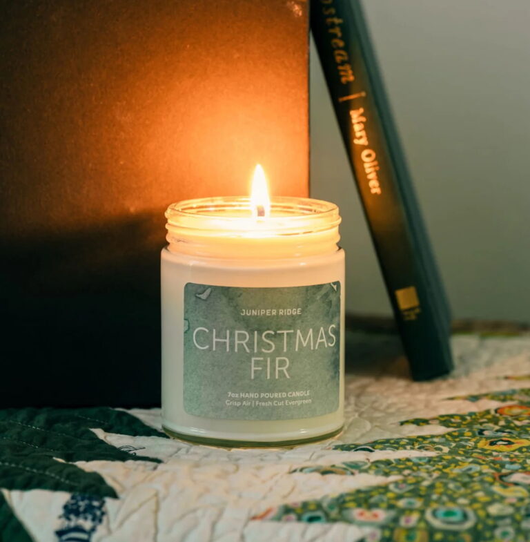 A Christmas Fir scented candle from the brand Juniper Ridge is lit and displayed next to a book and a pinecone.