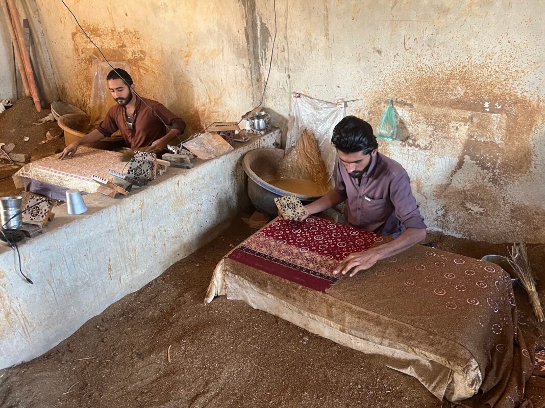 a man seated on the floor dios a block Ishaq had designed into dye and stamped the border of the fabric