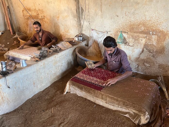 a man seated on the floor dios a block Ishaq had designed into dye and stamped the border of the fabric