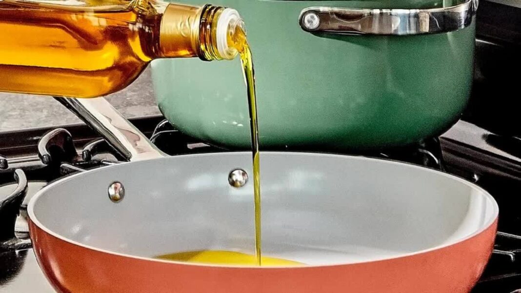 olive oil being poured from a clear glass bottle into a rust-colored frying pan on a stove, with a larger green pot behind it