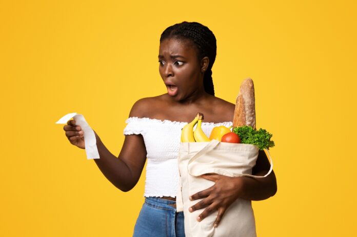 climate-flation Shocked young african american woman holding shopping bag and checking grocery receipt, emotionally reacting to big prices for organic healthy food, standing with open mouth over yellow background