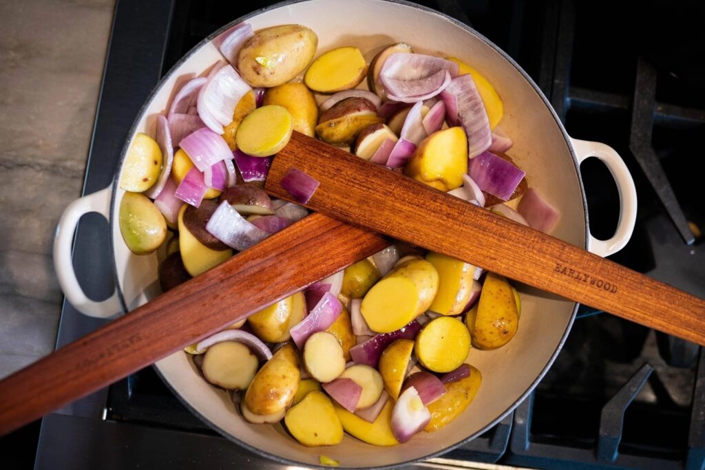Two wooden spatulas with the brand name Earlywood on them sitting on a cast iron enamel skillet of potatoes and onions