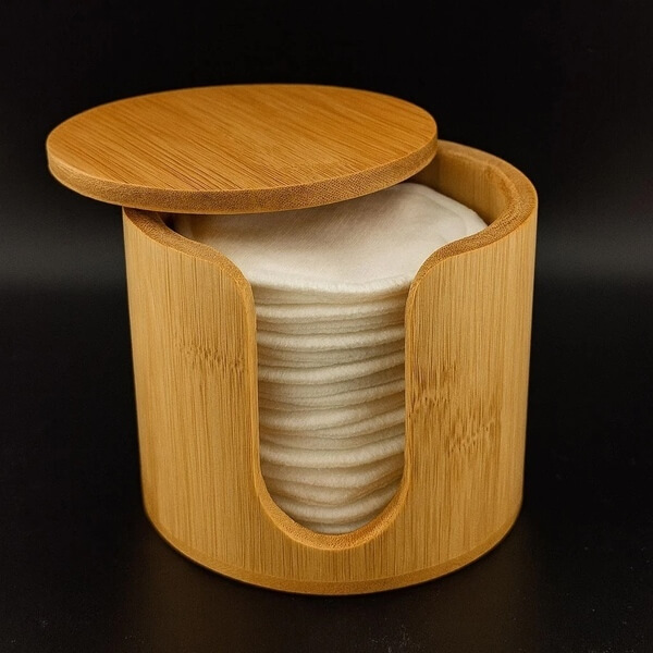 A bamboo storage canister with a sliding lid holds a neat stack of reusable cotton rounds, shown against a dark background.