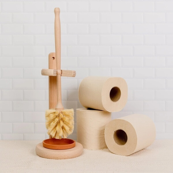 a toilet brush made of wood and natural materials next to three rolls of toilet paper